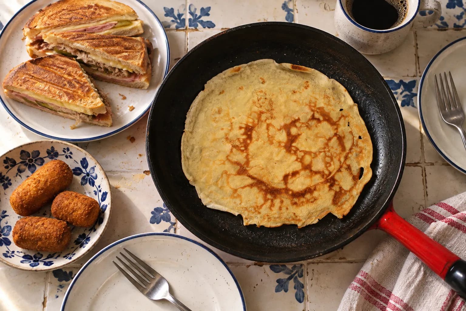 Overhead view of a shared breakfast: a thick pancake in a blackened cast iron skillet, a pressed sandwich cut on the diagonal and three small croquettes