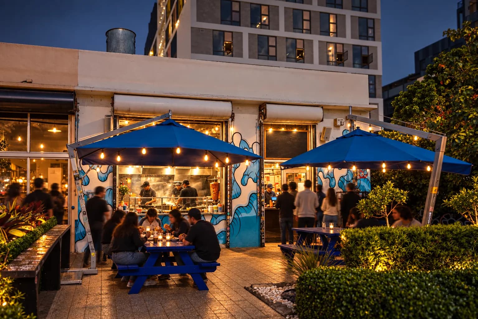 An open-fronted Wynwood taqueria at night with blue umbrellas over the forecourt tables and warm light spilling onto the pavement