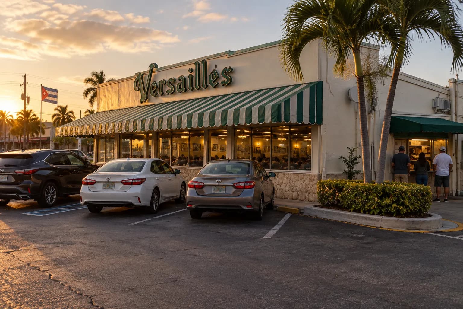 A low-rise Little Havana restaurant seen wide across its car park at dusk, interior lights on and a few people at the walk-up window on the side wall
