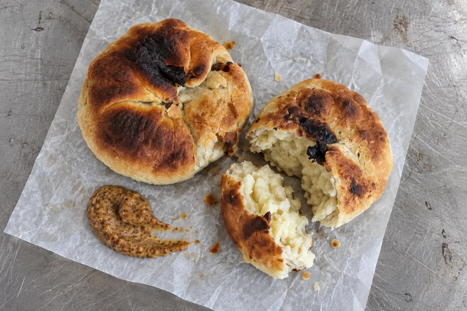 Two round baked potato dumplings on waxed paper on a worn steel counter, one cut open beside a smear of brown mustard, shot from directly above