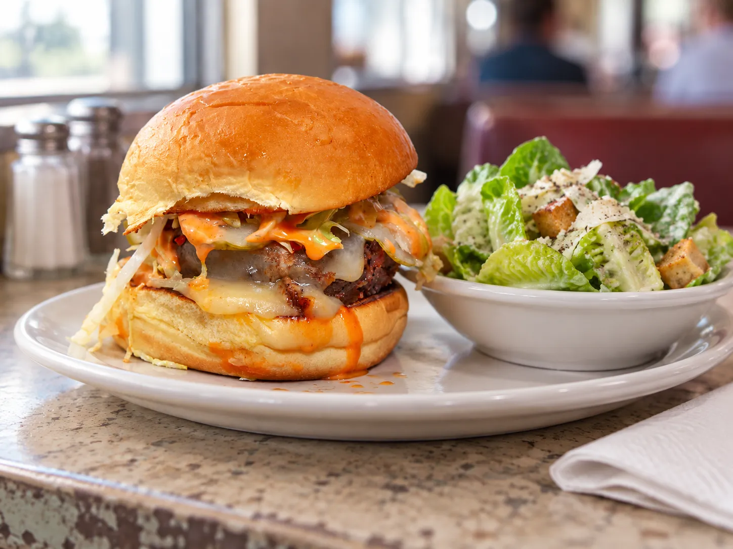 A vegetarian burger on a soft potato roll with pickles and shredded lettuce, plated on a diner-style counter beside a small dish of gem lettuce salad