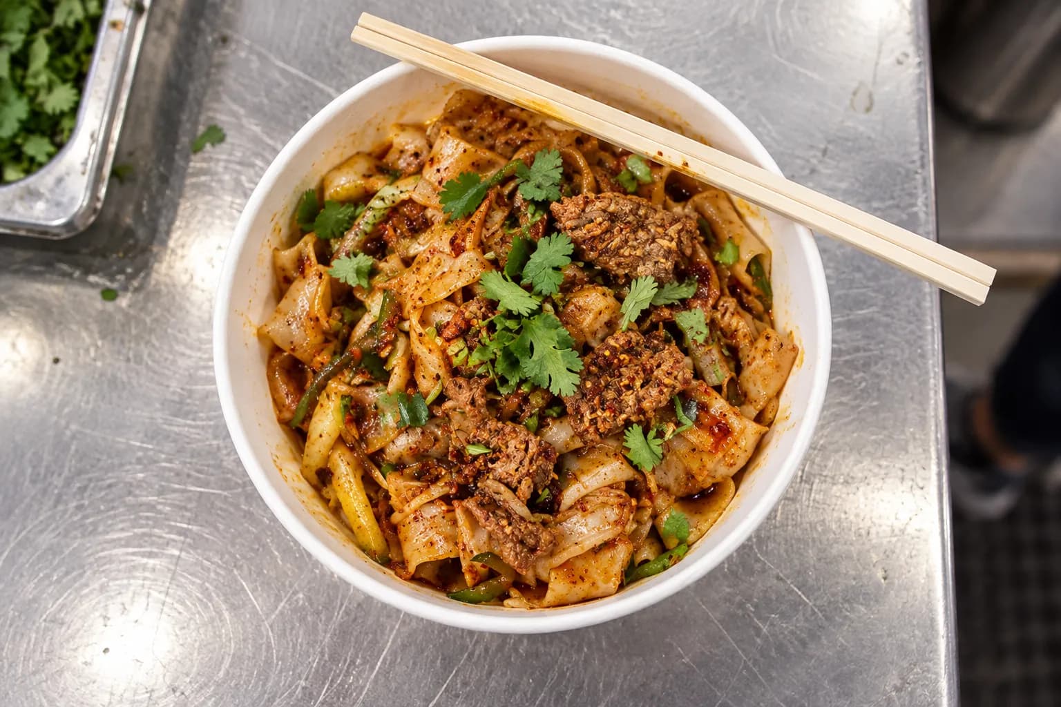 Overhead view of hand-ripped wide wheat noodles tossed with cumin-dusted lamb, chilli oil and coriander in a paper takeaway bowl on a stainless counter