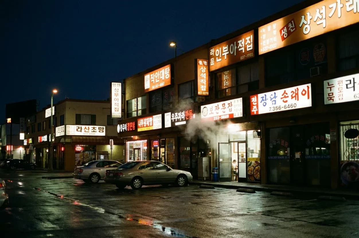A Koreatown Los Angeles commercial street after midnight, low-rise storefronts stacked with backlit signboards glowing amber and red, wet asphalt reflecting the light and steam drifting from a lit doorway