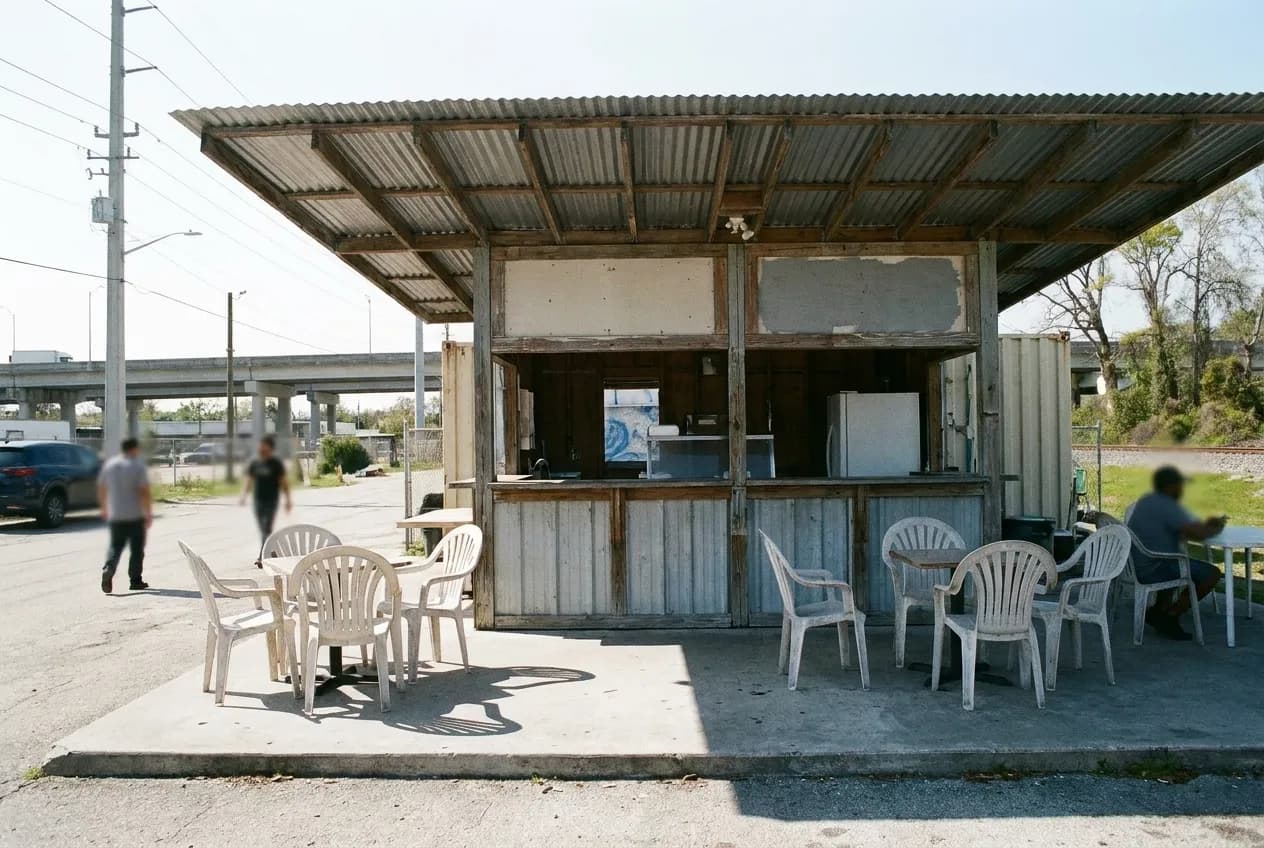 An open-sided food counter in bright Miami daylight, plastic chairs on a concrete apron and a corrugated roof throwing hard-edged shade