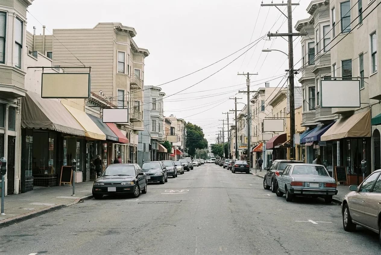 A flat commercial street of small shopfronts with layered awnings and hanging signage frames under overcast light, parked cars along both kerbs