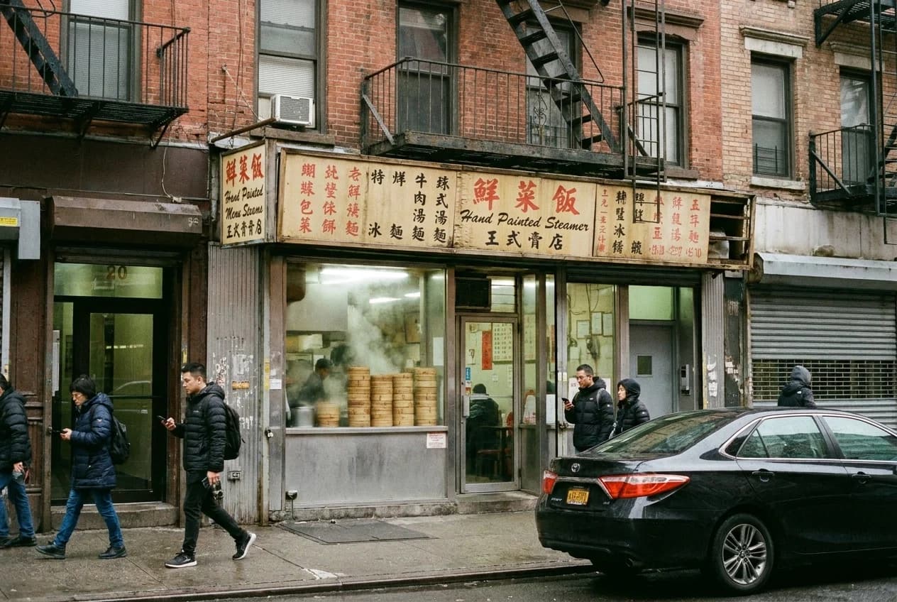 Narrow dumpling counter storefront on Eldridge Street with steamer baskets and hand-lettered menu boards