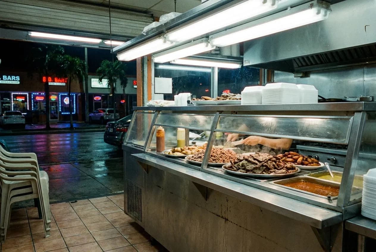 Steam tables piled with grilled meats and sides under bright fluorescent lights inside a Miami fritanga, with tiled floors and menu boards visible behind the counter.