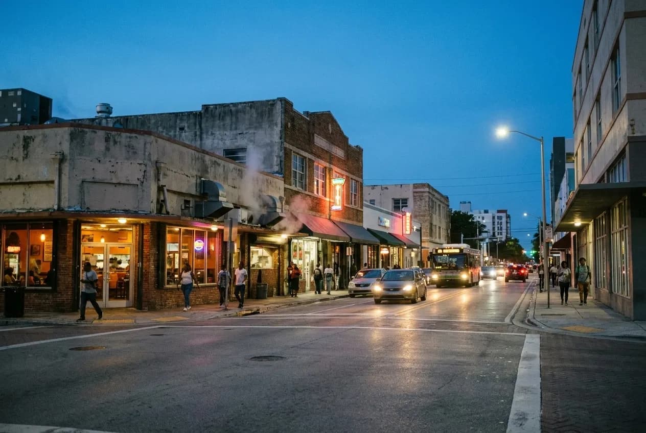 Wide view of a historic Overtown street at dusk with low-rise buildings, glowing storefronts, passing cars, and warm light spilling onto sidewalks under a deepening blue sky.