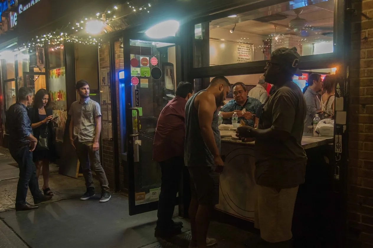 Corner slice shop glowing at night in Greenwich Village with people eating pizza at the open sidewalk counter