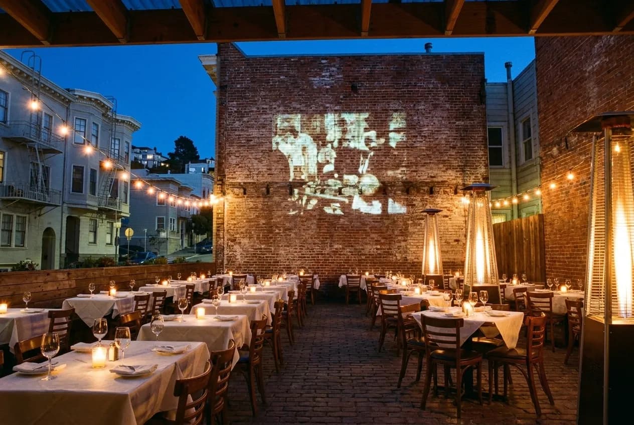 Outdoor courtyard tables at dusk with film projecting onto brick wall and string lights overhead.