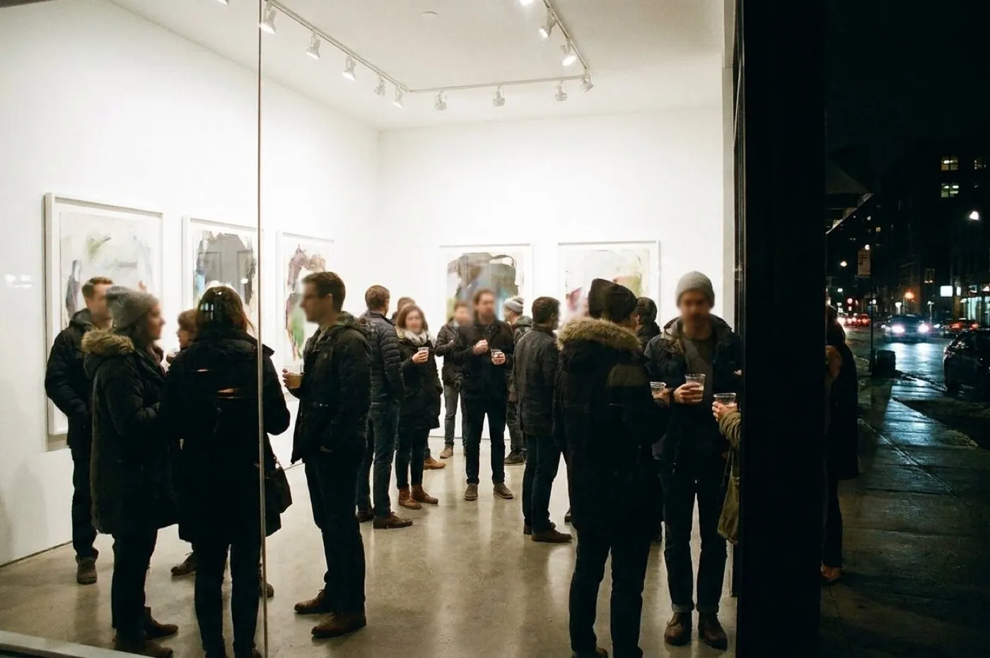 A gallery opening at night with people standing in front of large works, drinks in hand, the room bright against a dark street outside