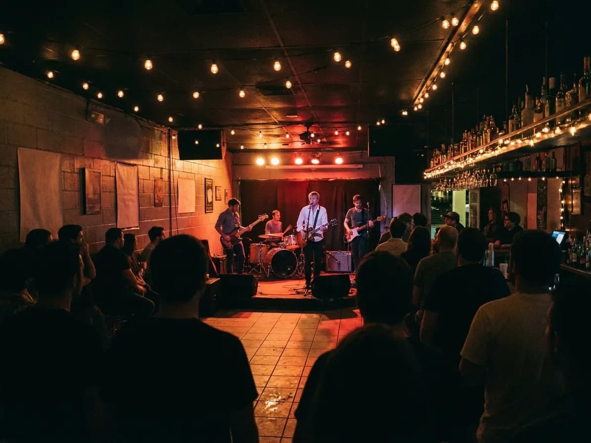 A four-piece band on a low stage in a small Miami dive bar, warm red light on breeze-block walls and a scattered crowd shot from behind