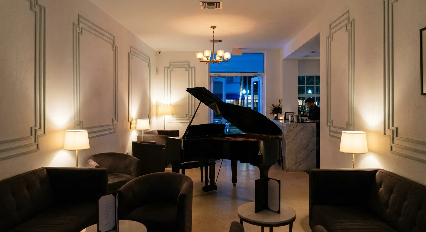 A grand piano under low light in the art deco piano bar of a small South Beach hotel, warm lamplight against a blue night doorway