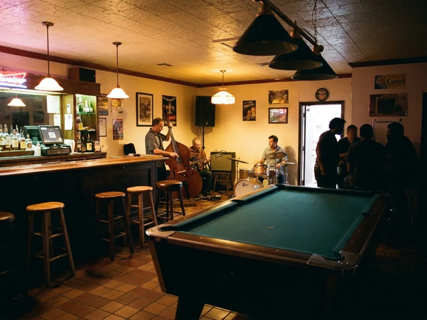 A small bar interior at night with a band setting up in the corner, mismatched stools, a lit back bar and a few early drinkers
