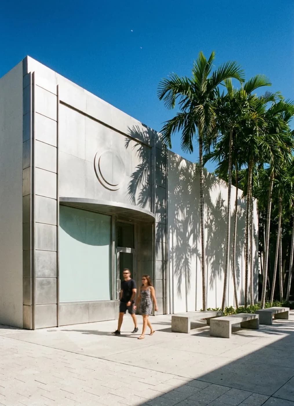 A modern museum courtyard with a sculptural metal facade, palms and concrete benches under bright sky