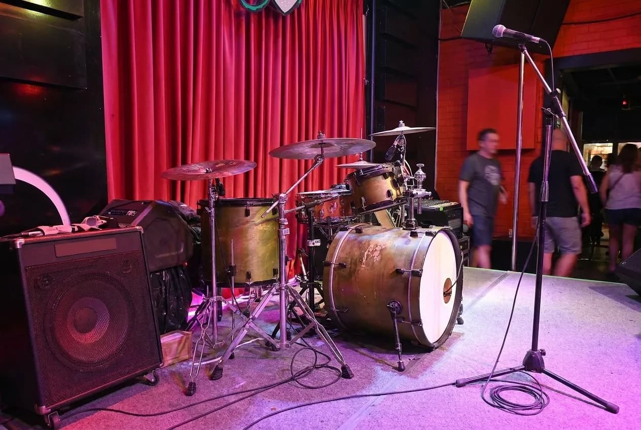 A small bar stage with a drum kit and two microphone stands under warm lights
