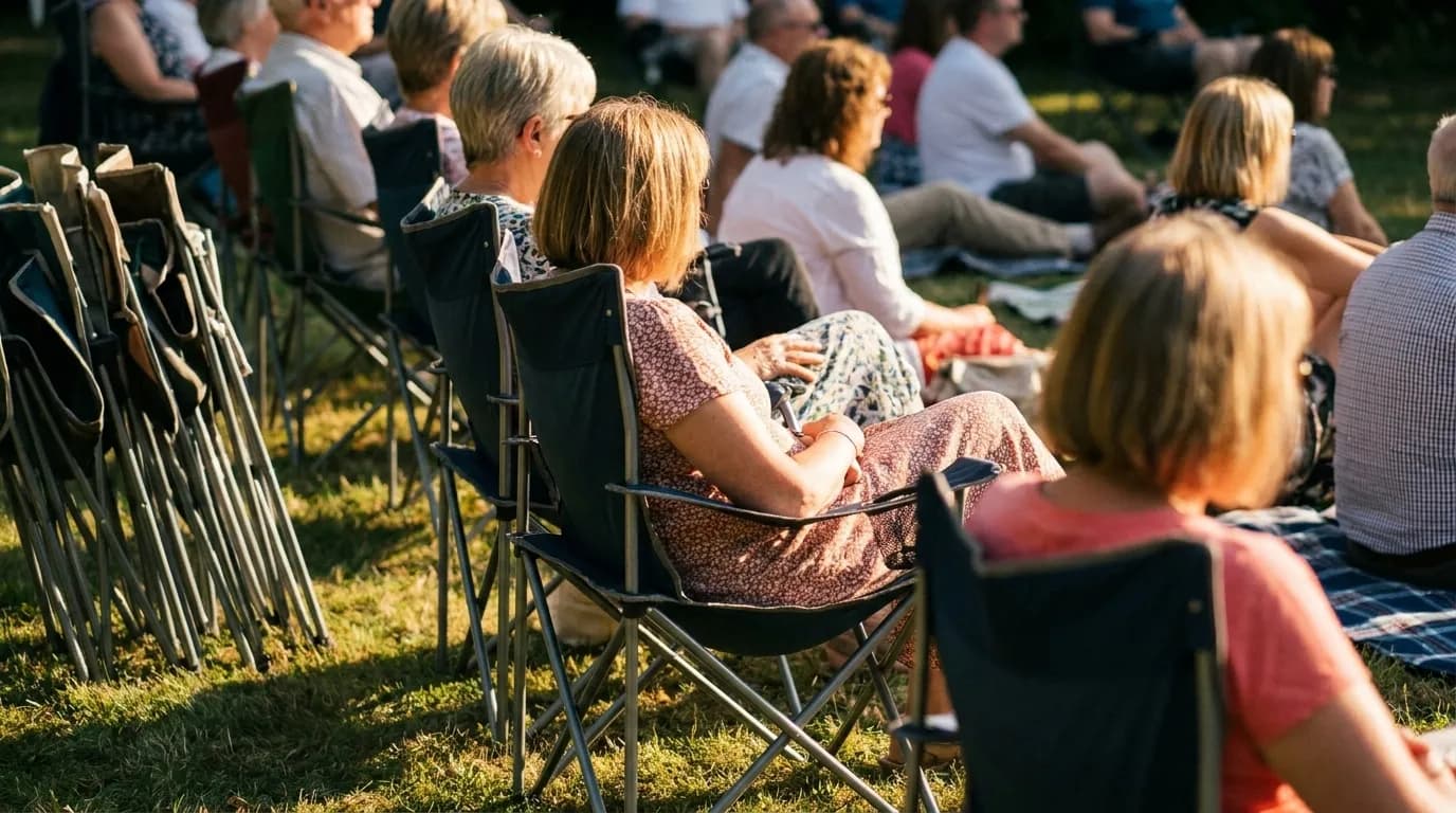 A close view of people seated on folding chairs and blankets on grass at an outdoor concert, a stack of stacked chairs to one side and warm stage light spilling across the crowd's shoulders