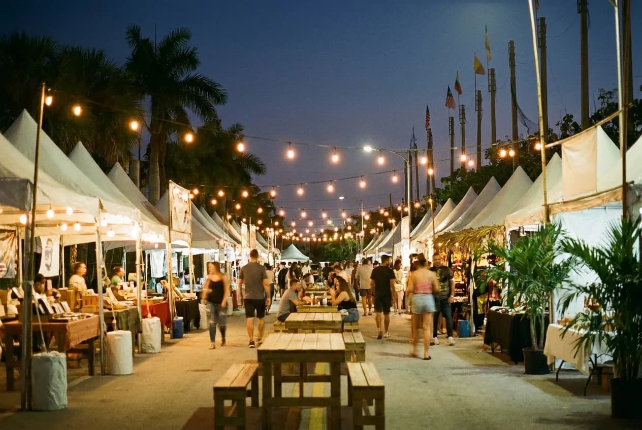 A closed street lined with canopied stalls in the evening, strings of bulbs overhead and people moving between the tables in warm light