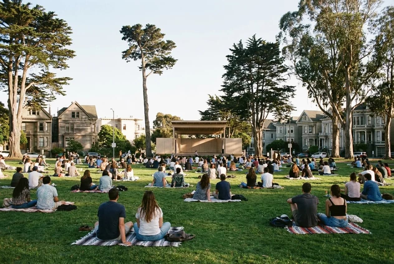 People spread across a broad park lawn on blankets facing away toward a distant stage, tall trees closing in on both sides