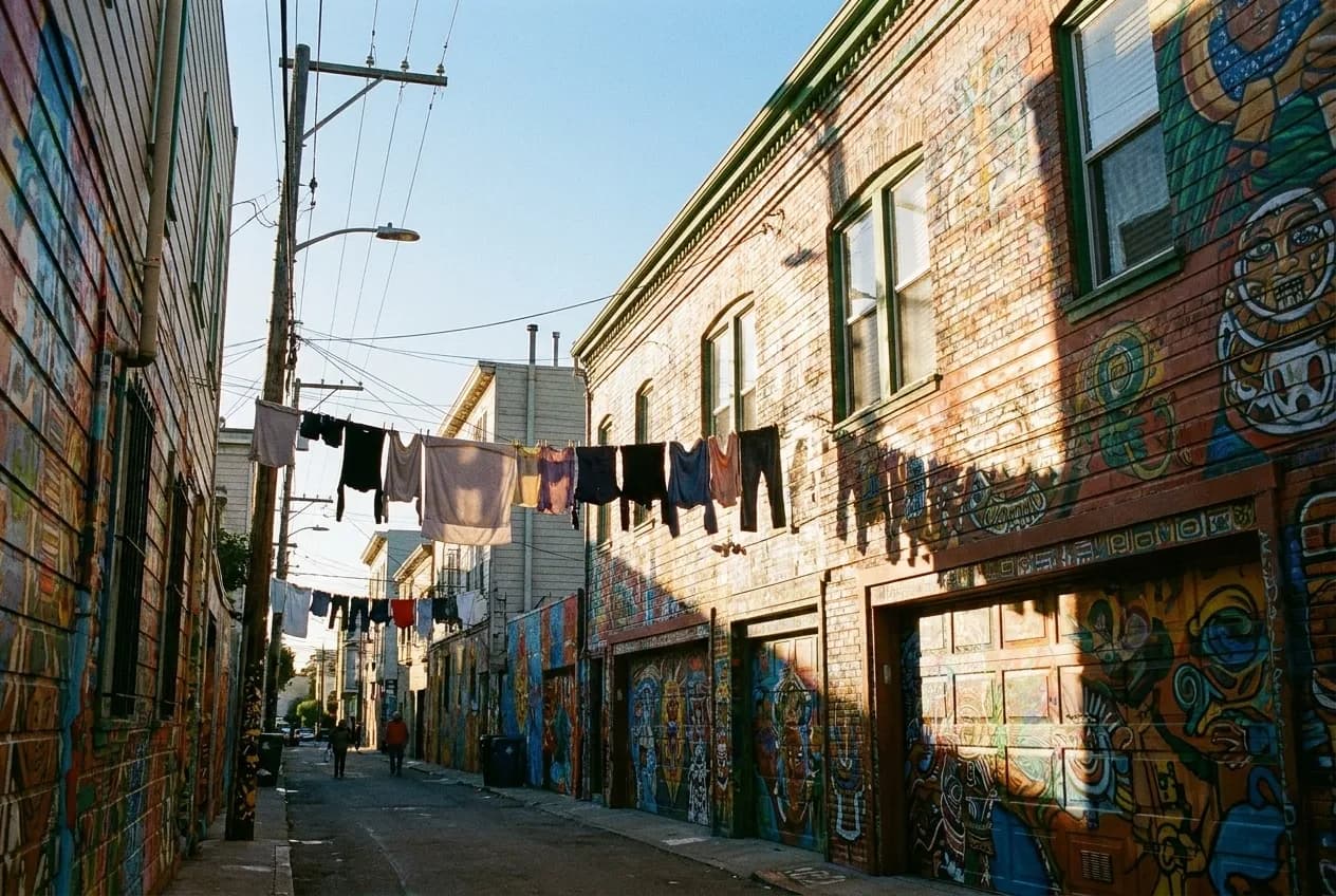 A narrow alley with both flanking walls and every garage door painted in dense murals, sunlight falling down one side and washing lines above