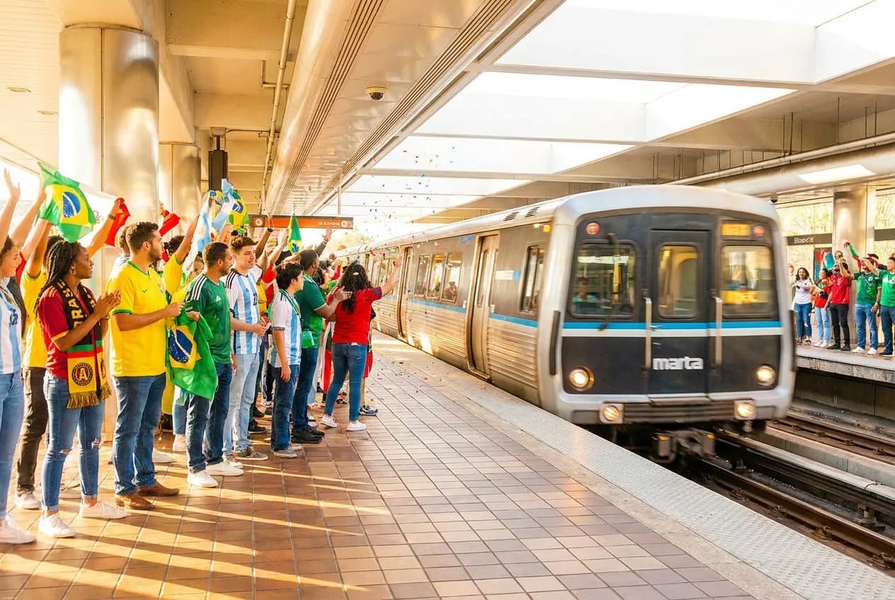A bright MARTA train pulling into a sunny Atlanta platform with fans in colorful jerseys and scarves ready for the World Cup semifinal