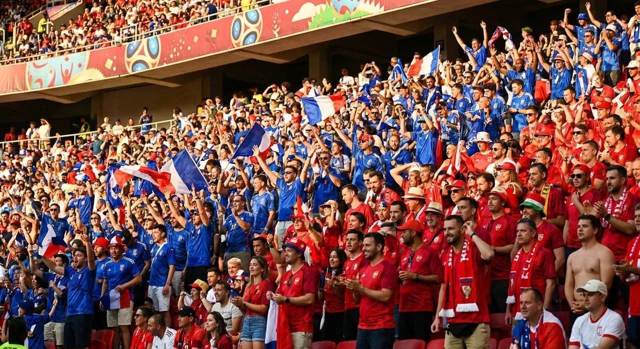 Enthusiastic fans wearing blue and red scarves celebrating in a sunlit stadium stand with flags waving