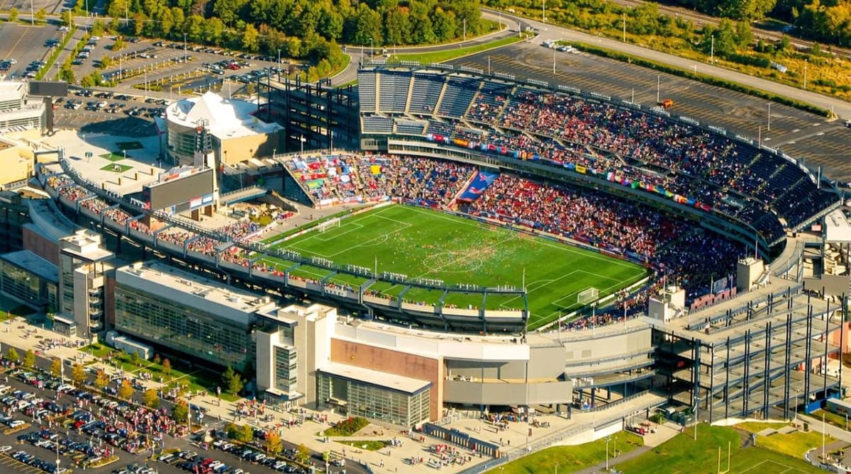 A large open-air stadium in a green New England setting under bright sky, calm after a big match
