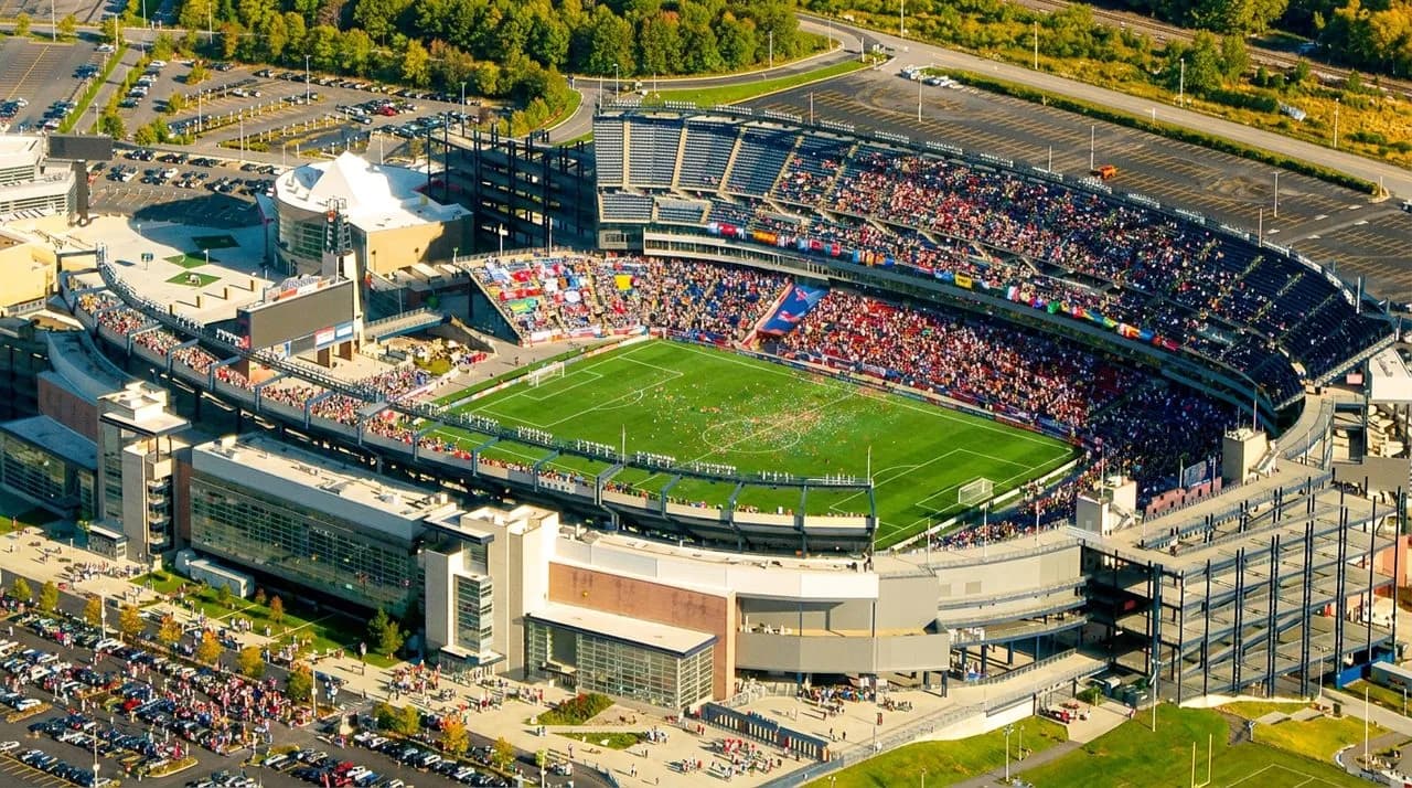 A large open-air stadium in a green New England setting under bright sky, calm after a big match