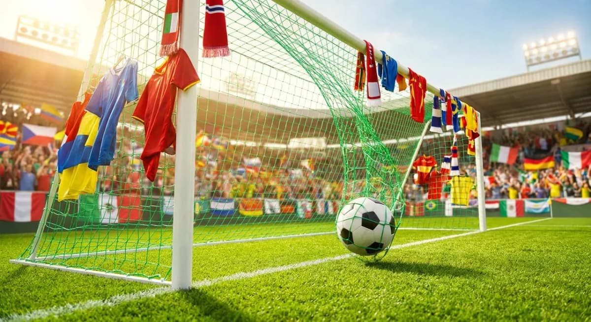 A football crossing the goal line under bright stadium floodlights with vivid green pitch and colorful crowd accents in warm natural light