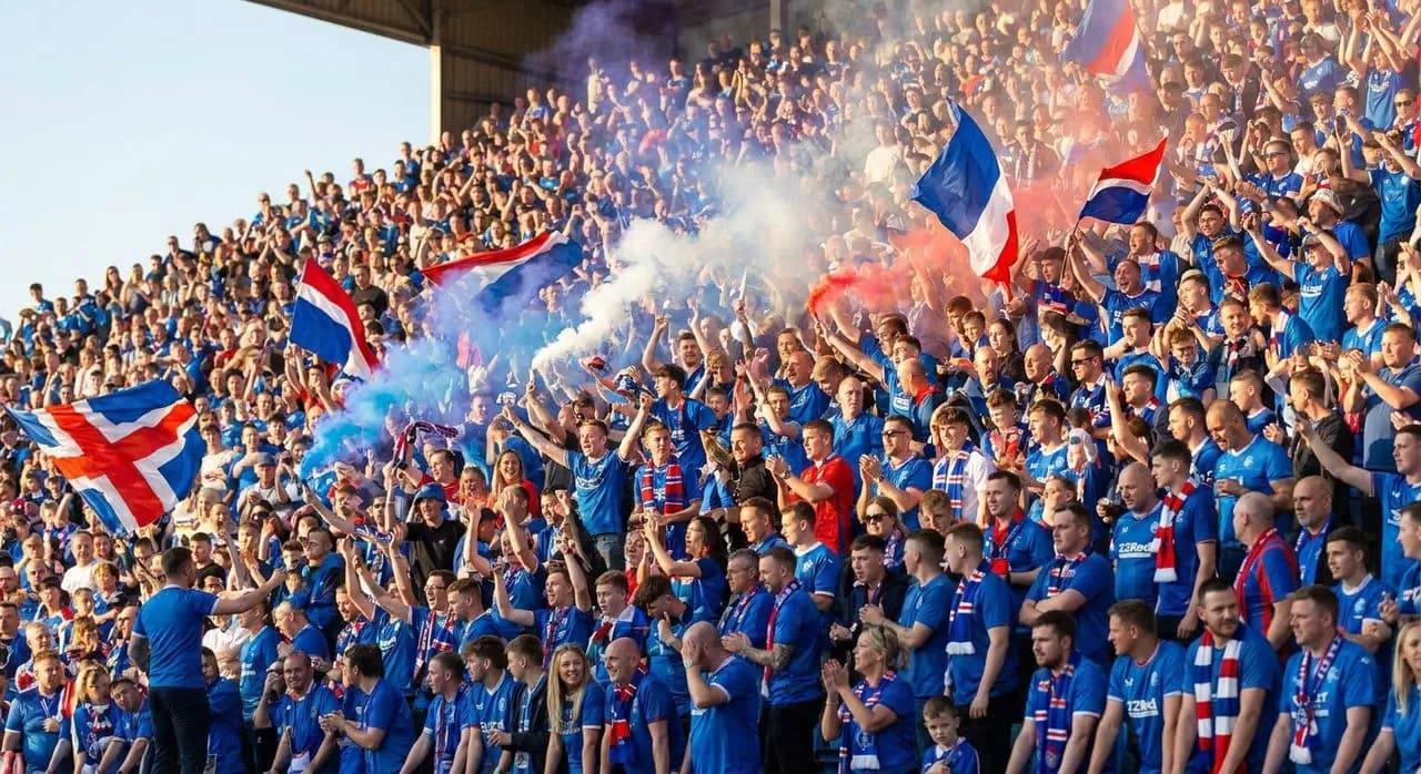 Fans wearing blue France jerseys and face paint celebrating in a sunny stadium stand, waving flags under bright afternoon light