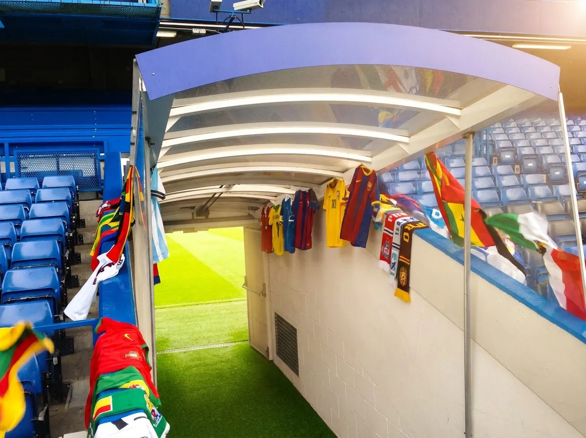 A bright stadium tunnel opening onto a vivid green pitch, bathed in warm natural light with festive jerseys visible along the walls