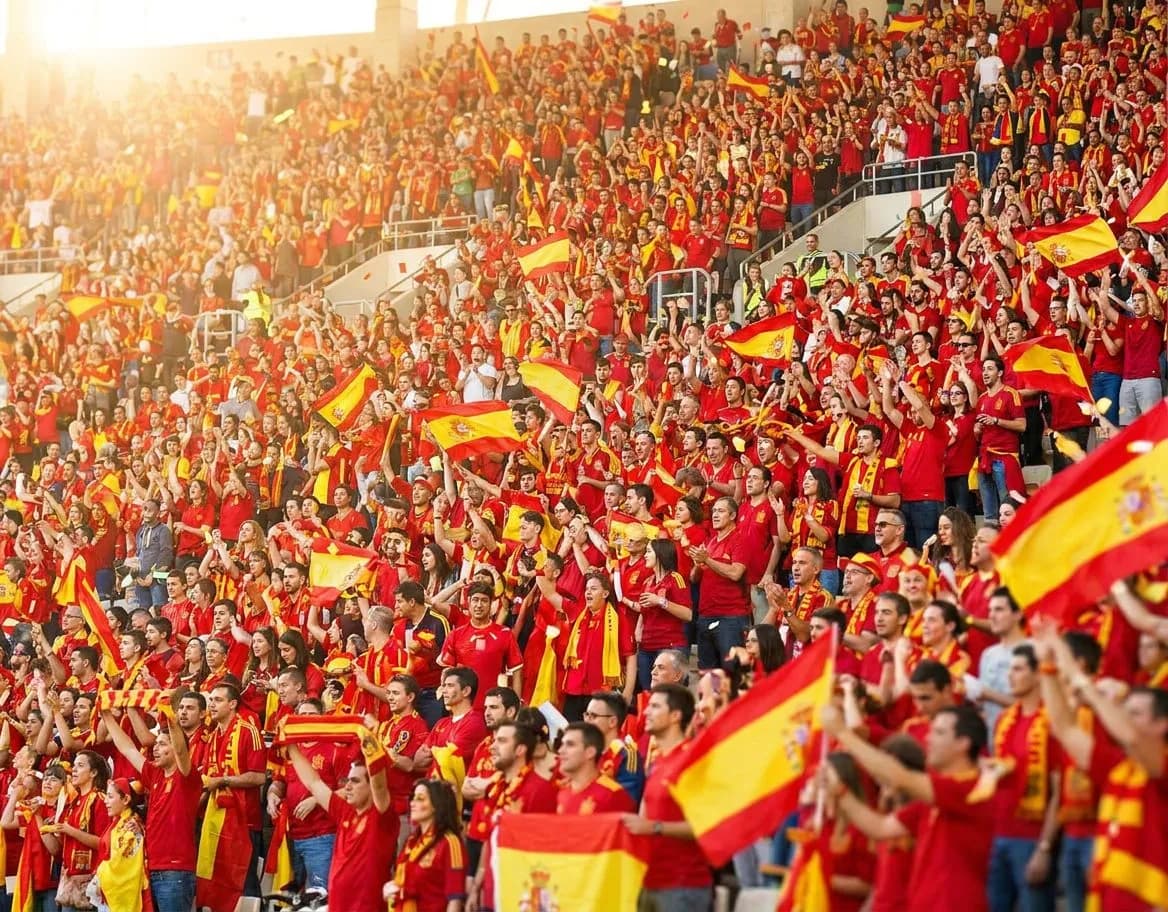 Spanish and Belgian fans in red and yellow-red jerseys mingling together in a sunny stadium stand, celebrating the festive spirit of the tournament.