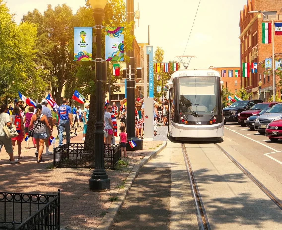 A bright Kansas City streetcar gliding down a sunny downtown street adorned with colorful banners, fans in jerseys and scarves waiting at the stop under clear blue skies