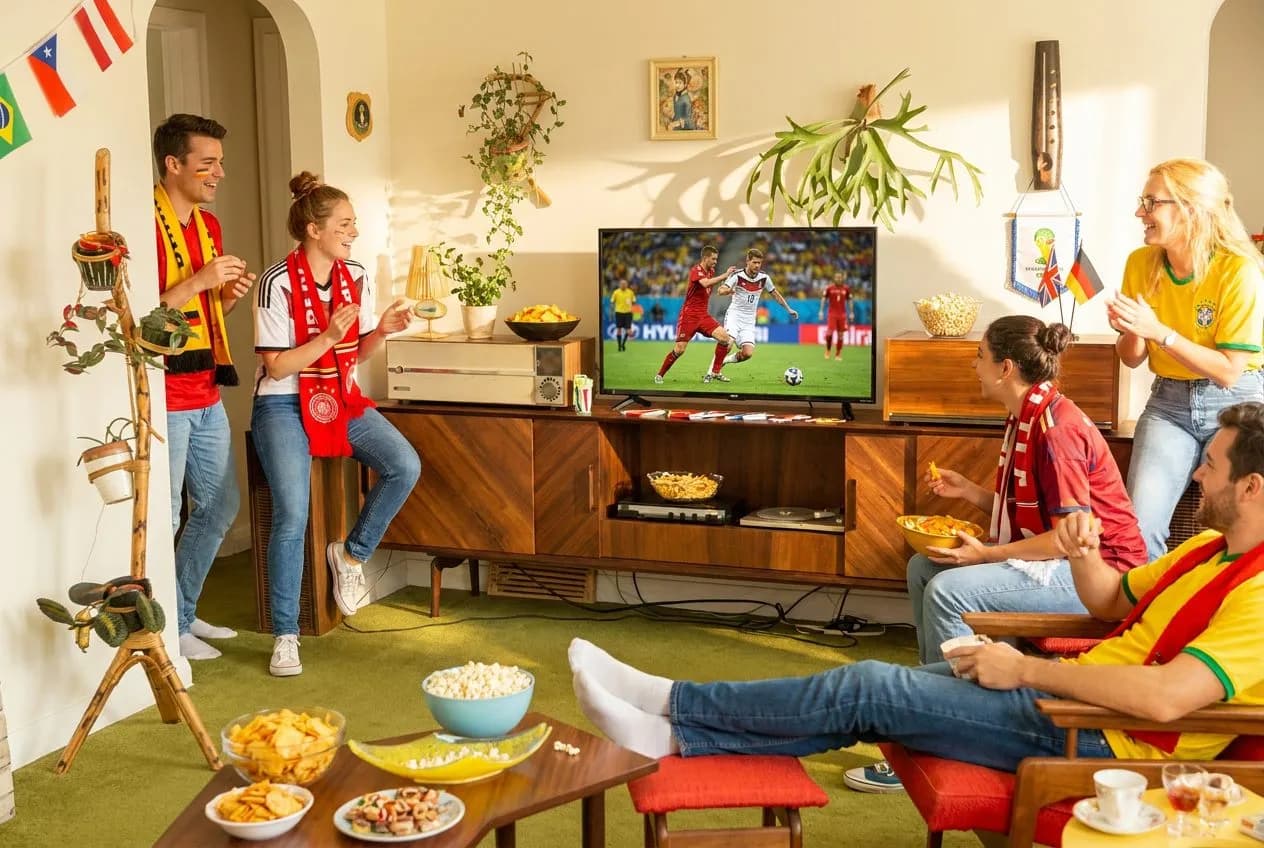 A bright living room with a large screen showing a World Cup match, friends gathered with snacks, jerseys and scarves visible, warm natural light streaming through windows