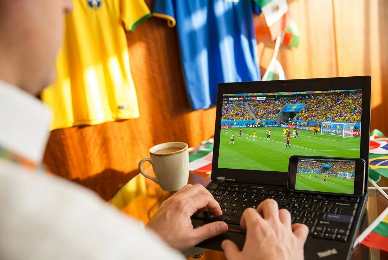 A laptop and phone on a wooden table streaming a live match beside a coffee cup, sunlight casting warm shadows across the setup