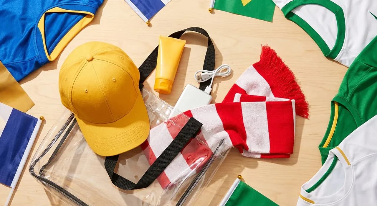 Flat-lay of World Cup match-day essentials including clear stadium bag, baseball cap, sunscreen tube, portable charger, and colorful team scarf on bright surface