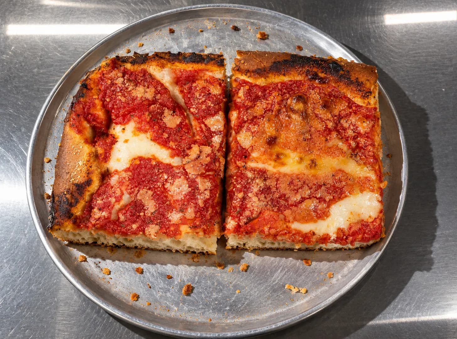 Thick square pizza slices cut from a sheet pan with a deep airy crumb at the cut edge and heavy browned cheese, on a crumb-dusted metal tray