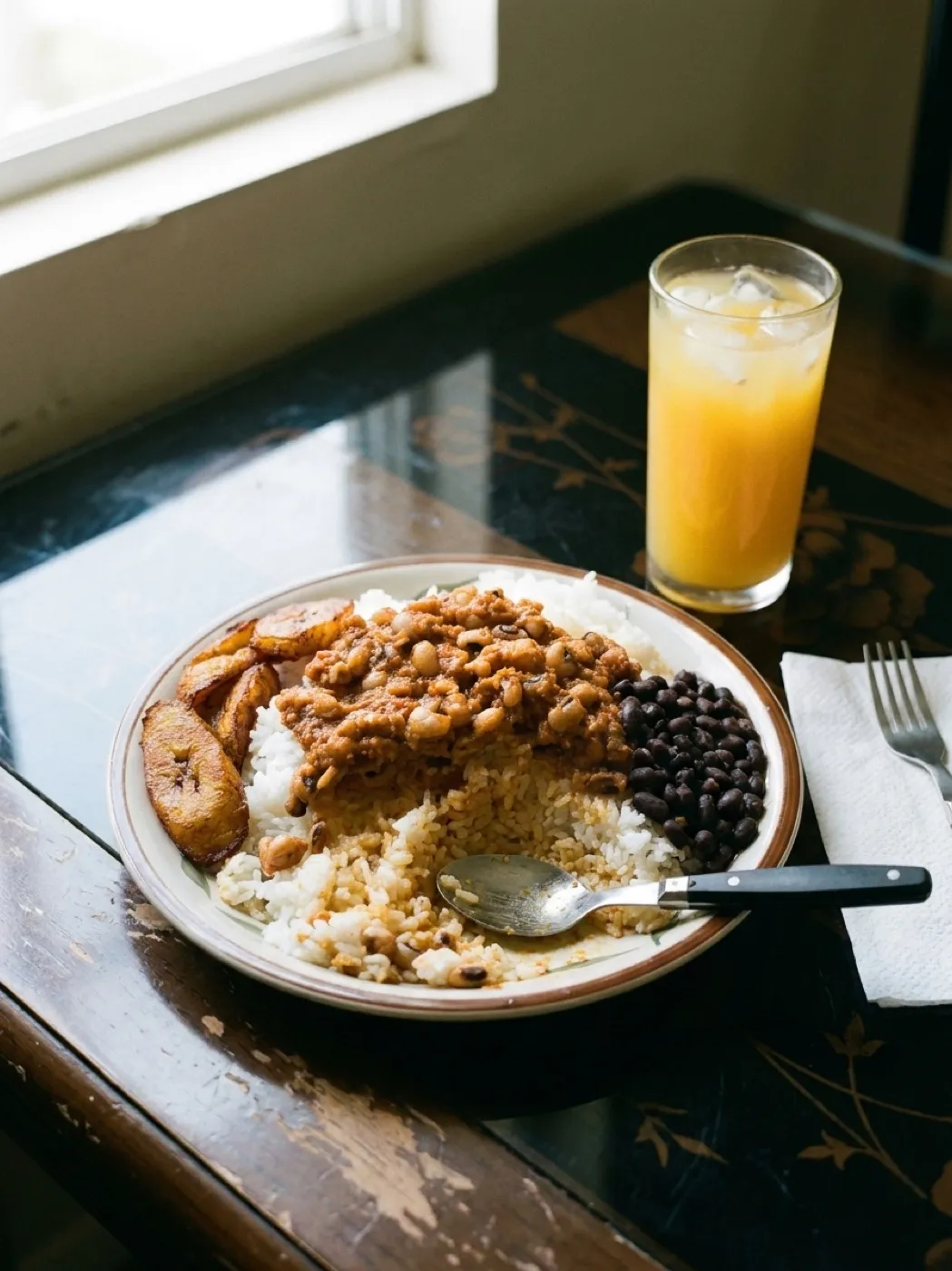 A plate of oxtail stew with white rice, black beans and fried sweet plantains on a laminate table beside a glass of iced juice