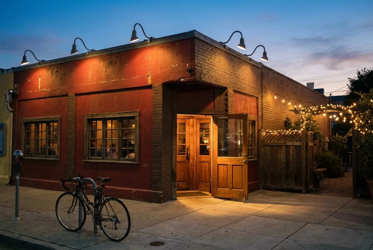A neighbourhood bar front at dusk with warm light spilling onto the pavement and string lights over a side gate