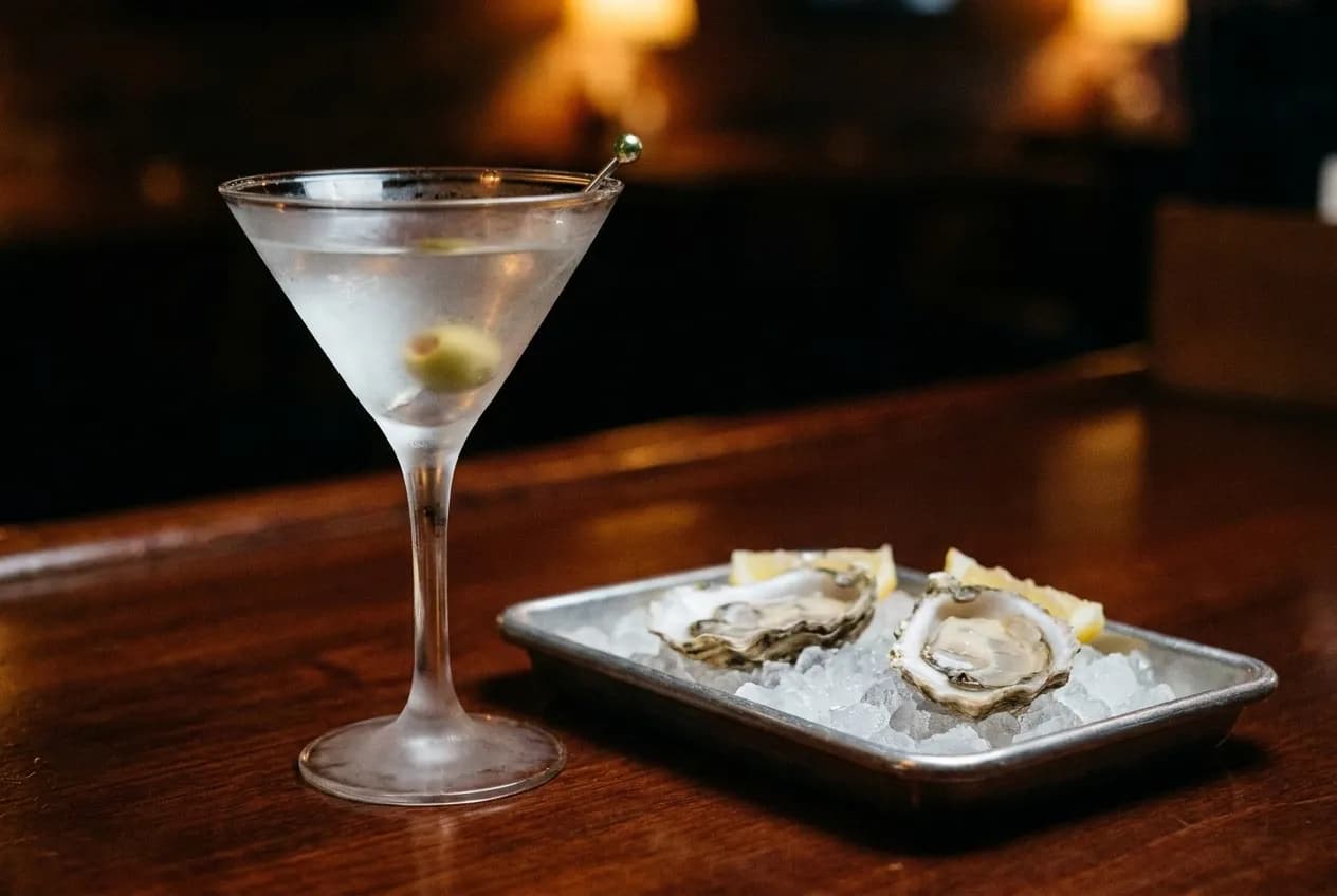 A chilled martini glass beside two open oysters on crushed ice on a dark polished bar under low warm light