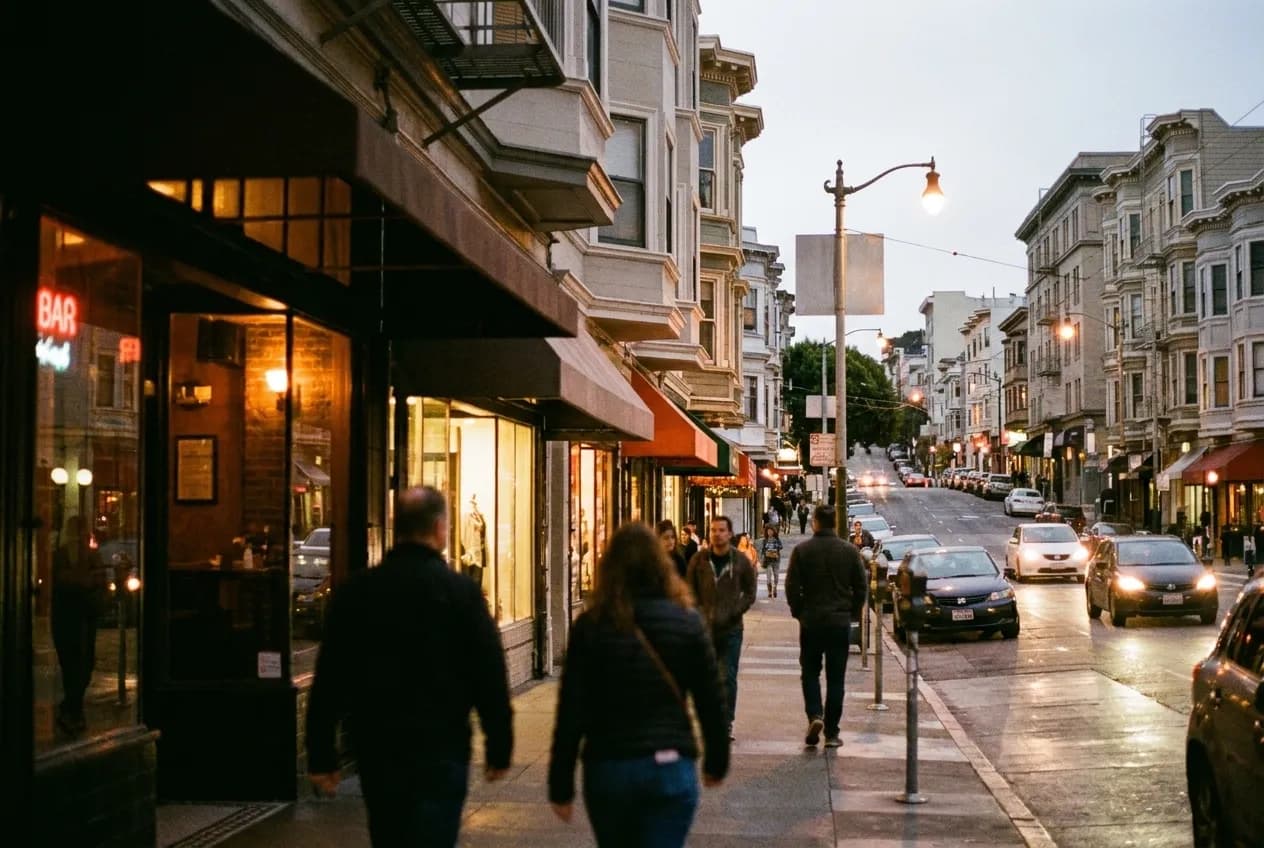 A San Francisco commercial street in early evening with bar windows lit and people on the pavement