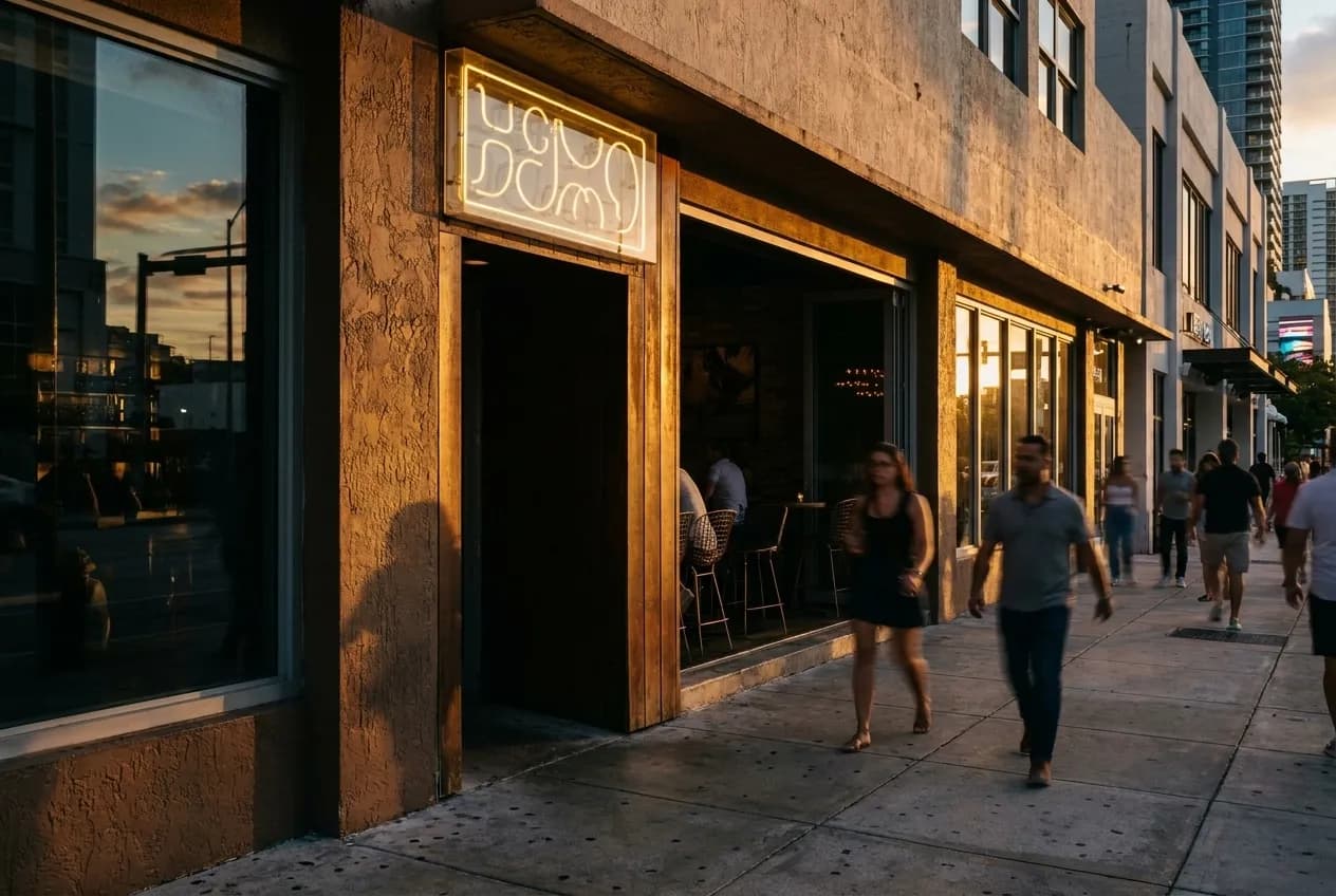 A compact Downtown Miami bar frontage at street level, neon glowing softly in early evening as pedestrians pass under awnings on the sidewalk.