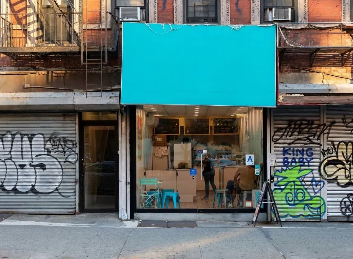Turquoise-awning ice cream shop storefront between graffiti-covered shuttered gates on a Lower East Side block