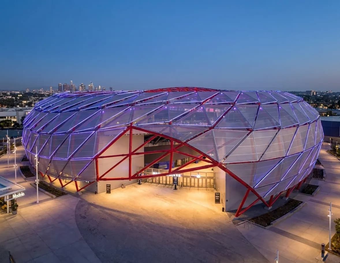 Night exterior view of Intuit Dome's distinctive curved architectural shell with illuminated facade and surrounding plaza area in Inglewood