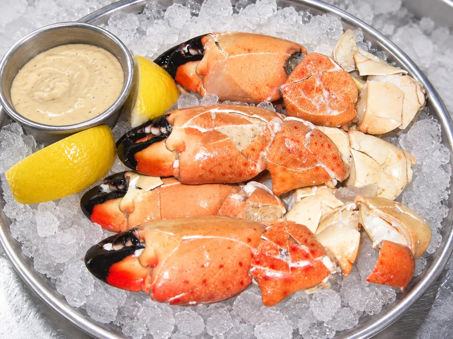 Chilled stone crab claws with cracked mottled orange and black shells arranged on crushed ice on a metal tray, with a ramekin of mustard sauce and lemon wedges