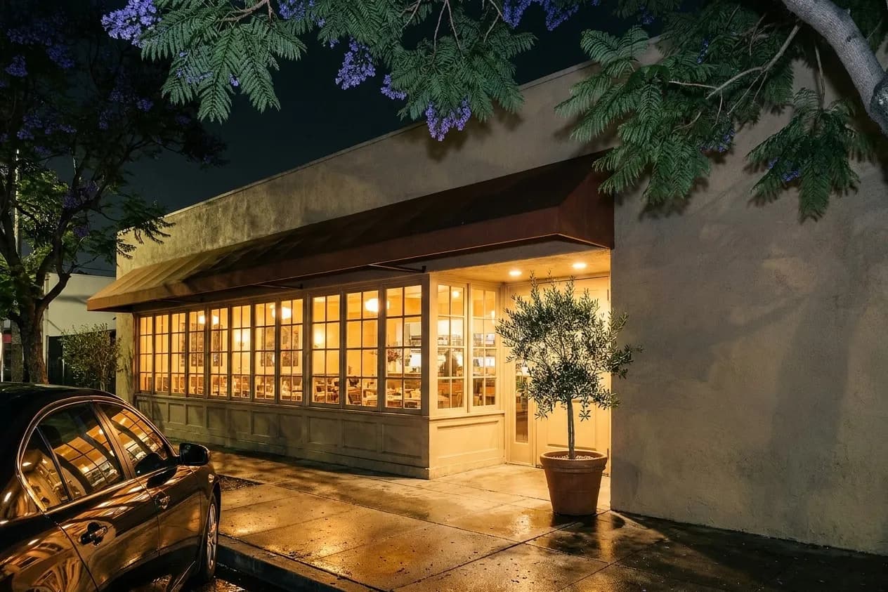 A low stucco Los Angeles restaurant frontage after dark, warm window light spilling onto the sidewalk beneath a deep awning