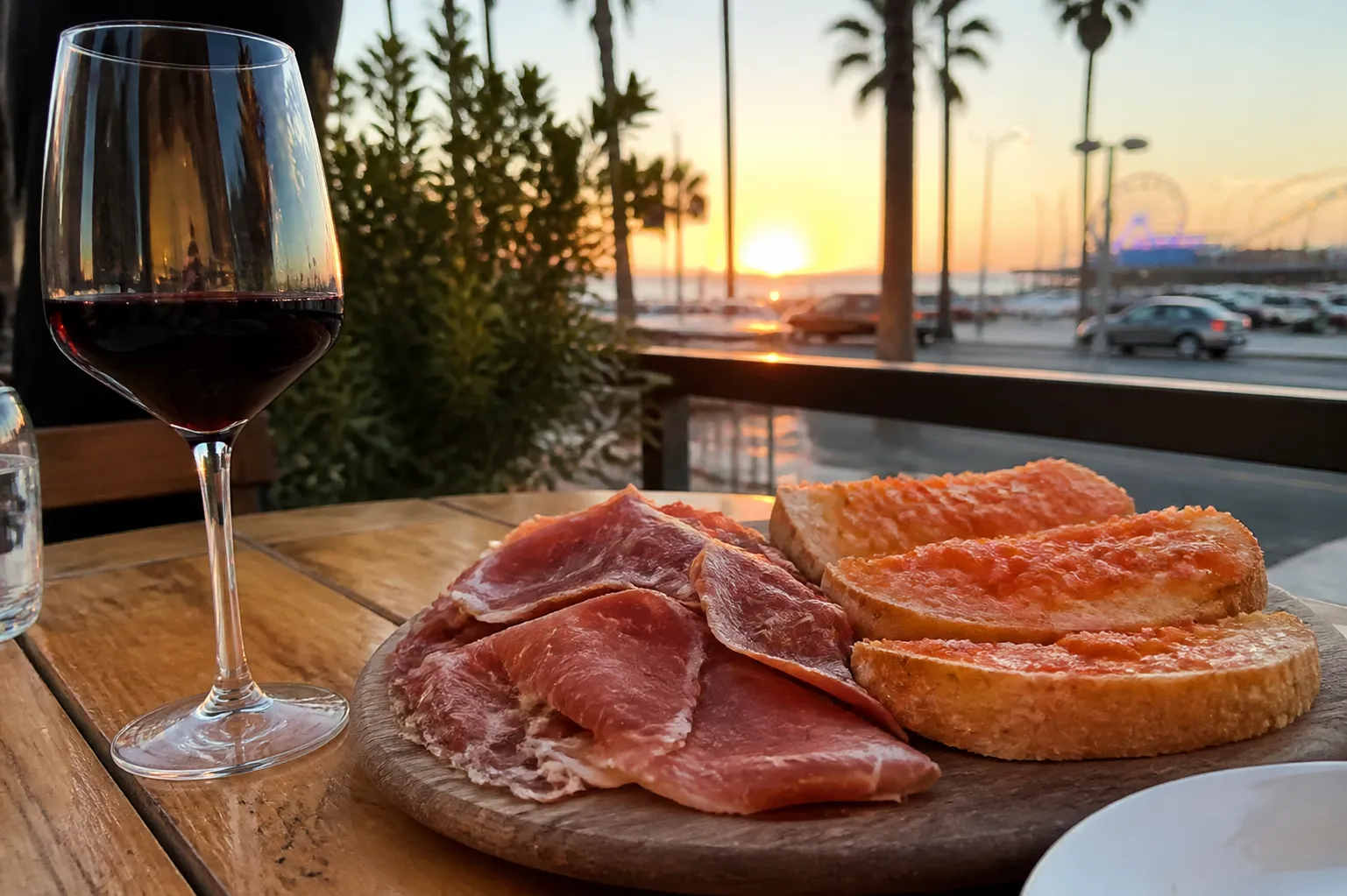 A close-up shot of a dark wood table filled with plates of pan con tomate, patatas bravas, and two glasses of Spanish Rioja at sunset.