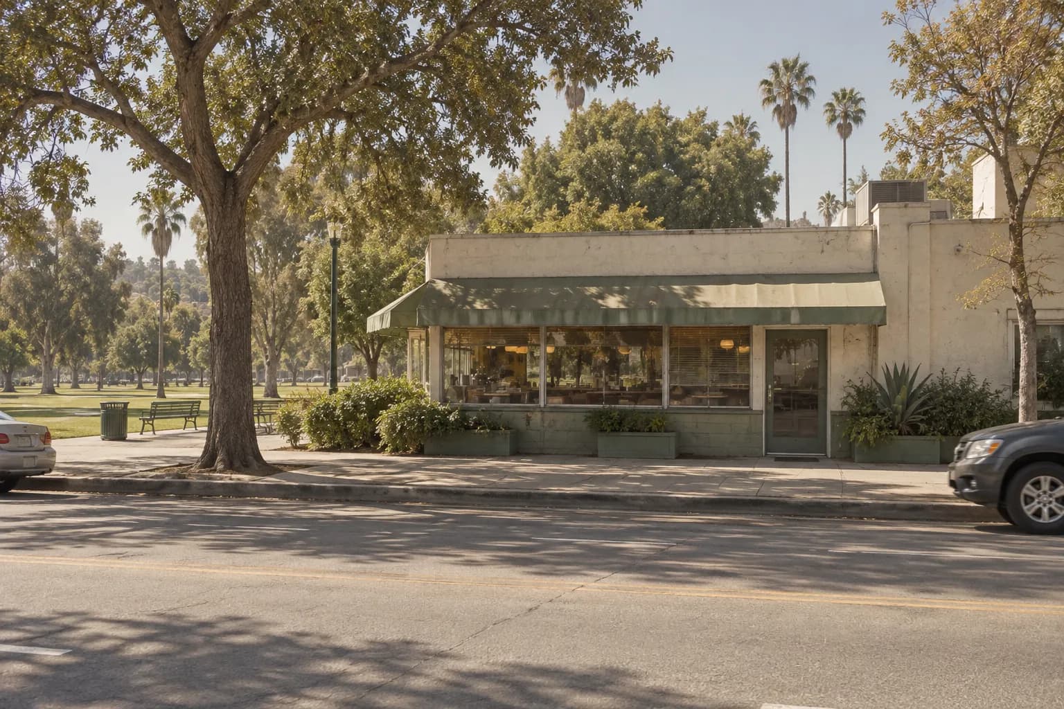 A daytime street view of a low-rise breakfast cafe across from a broad urban park, with North Spring Street, paving, mature trees and muted Los Angeles sunlight forming a documentary city scene.