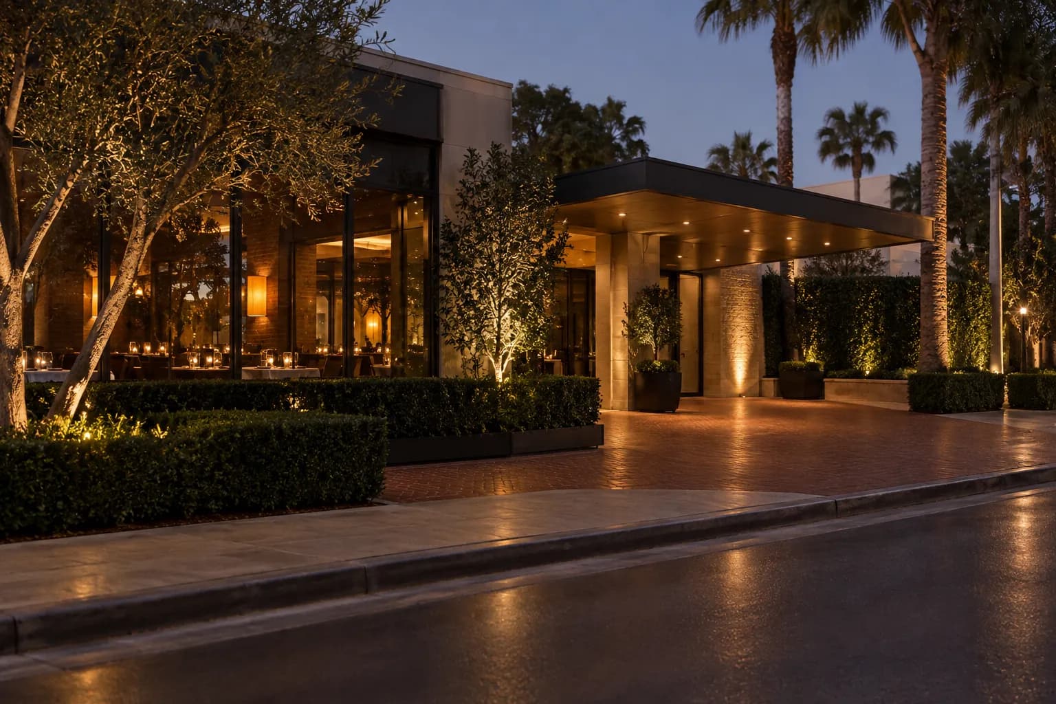 A wide evening view along a polished Beverly Hills street, with a low restaurant facade, leafy planting, warm interior light, brick paving and cars approaching a valet entrance.