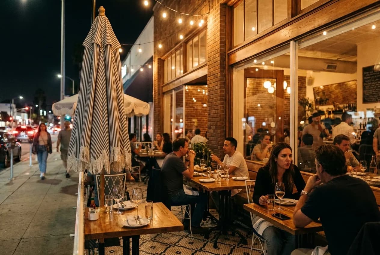 Night view of illuminated restaurant patio on Abbot Kinney Boulevard with diners seated outdoors under string lights and warm interior visible through windows